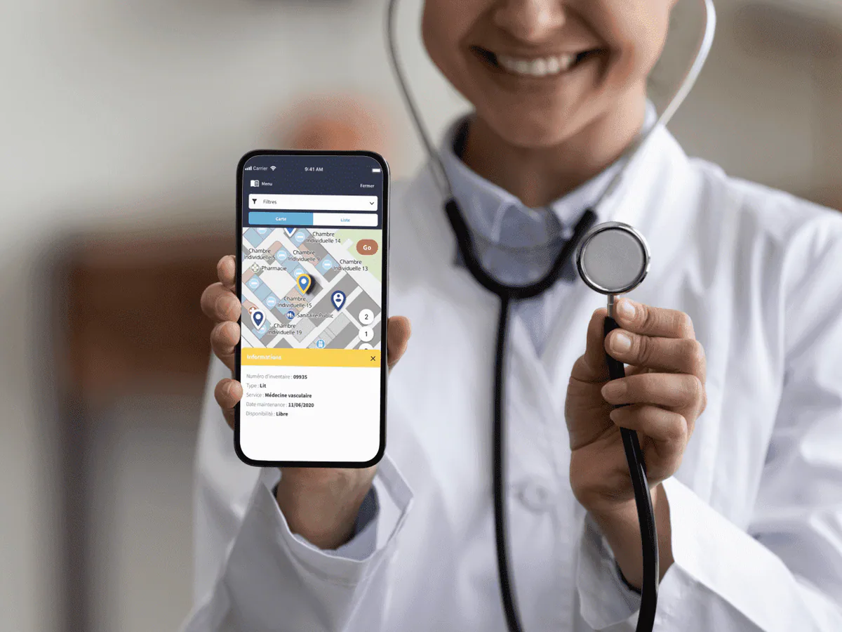 Une infirmière tient un smartphone affichant l'application Ubudu RTLS, avec le matériel médical localisé sur le plan d'un service et la fiche du lit sélectionné