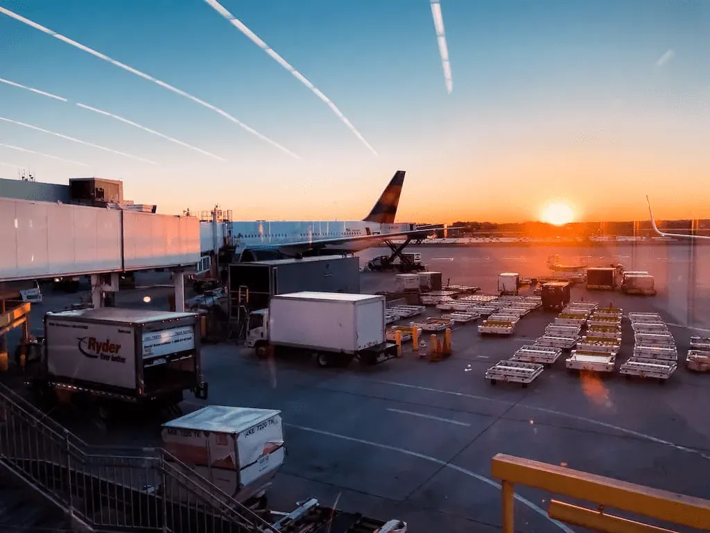 Placa de estacionamento do aeroporto ao pôr do sol, com uma aeronave na porta de embarque e carrinhos de bagagem, activos do lado ar que o RTLS pode rastrear
