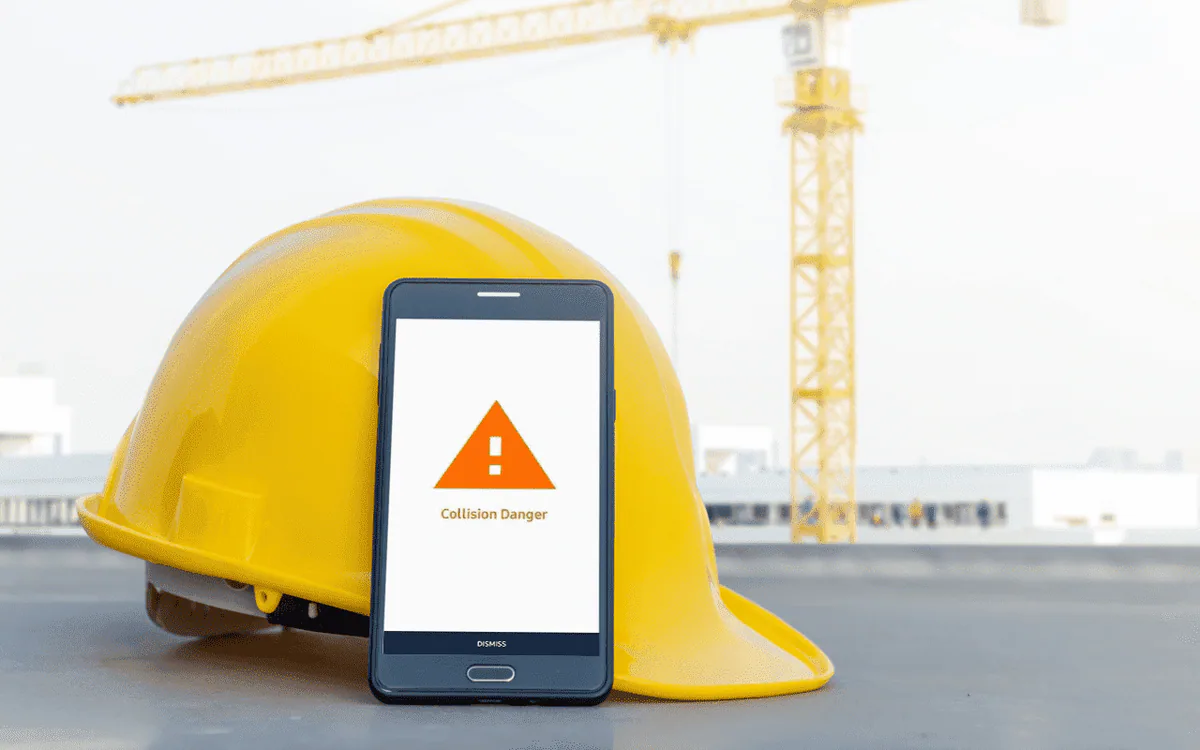 Um capacete de segurança ao lado de um smartphone com um alerta de «Perigo de colisão», com uma grua de construção ao fundo