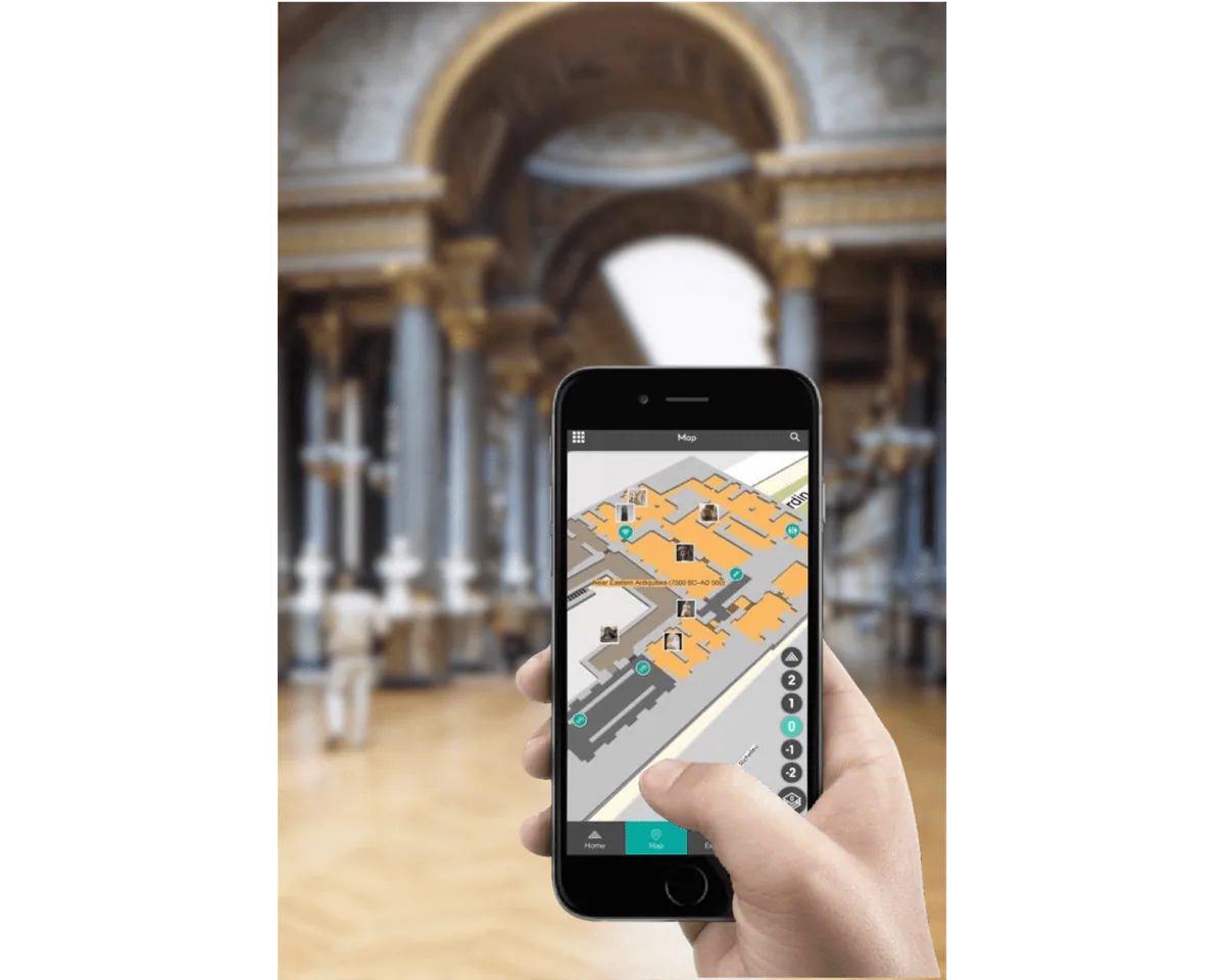 Una mano que sostiene un smartphone con el mapa interior de Ubudu de un museo, con la galería del recinto al fondo